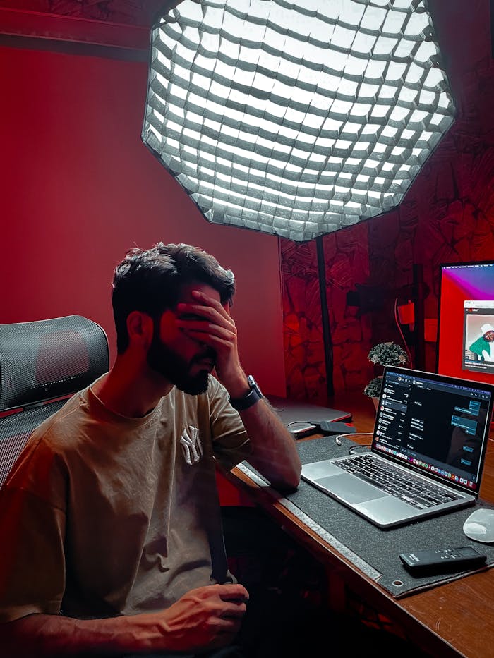 A tired man sits at a cluttered desk with a laptop under softbox lighting, looking stressed.