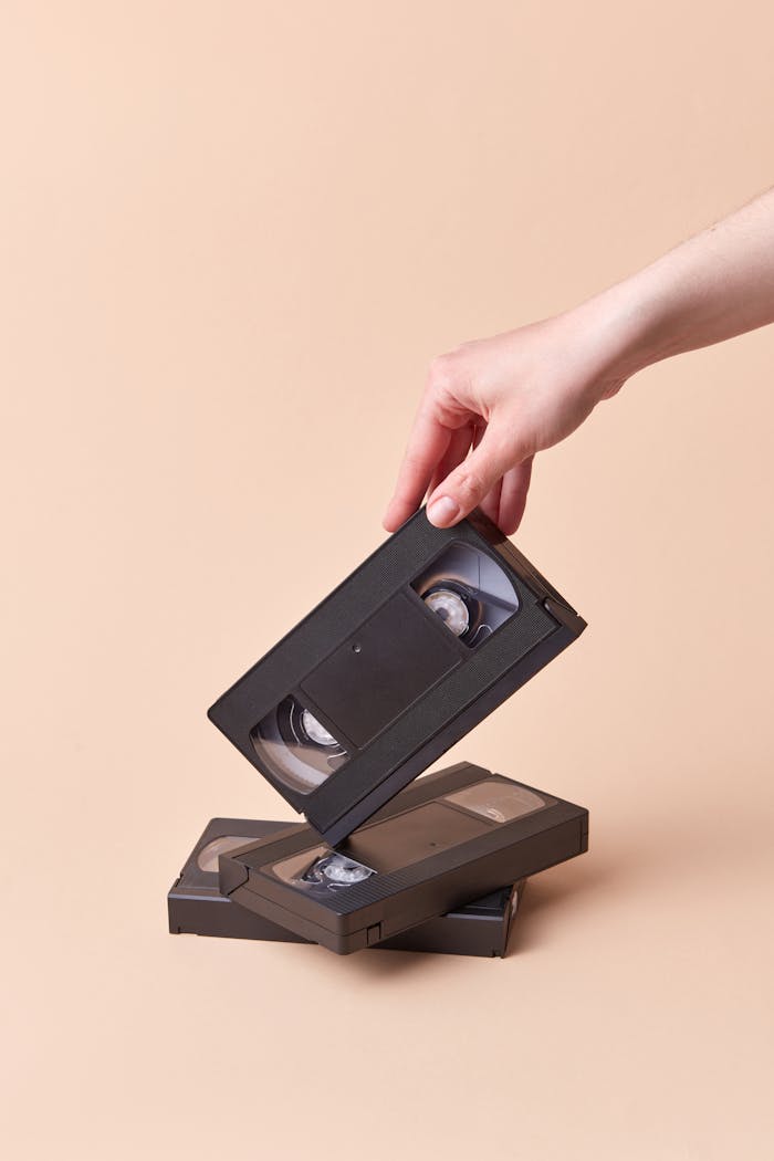 A close-up of a hand holding a stack of retro VHS tapes on a beige background, highlighting nostalgia.